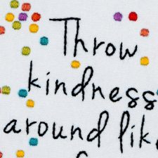 Close-up van geborduurde tekst 'Throw kindness around like confetti' met kleurrijke confettistippen.